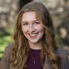 Bella Gray wears a burgundy shirt and a brown plaid blazer with a gold necklace. She smiles and stands in front of a blurred forest background.