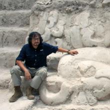 Carlos Morales-Aguilar wears a blue button-down shirt. grey pants, and brown boots. He sits on the steps of an enormous, ancient white stone structure, with ornate curved carvings on the walls.