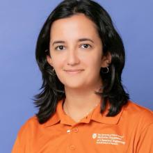 Lea El Khoury wears an orange polo with text on the left side of the chest reading "The University of Texas at Austin McKetta Department of Chemical Engineering, Cockrell School of Engineering." She stands in front of a light indigo background.