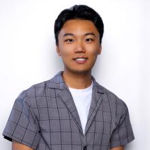 Cheng Chow wears a grey button-down shirt and stands smiling in front of a white background.