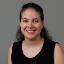 Kate Windsor wears a sleeveless black shirt and black hoop earrings and smiles in front of a grey background.