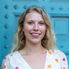 Erin Crownover smiles in front of a bright teal door. She wears a white v-neck dress printed with orange and purple leaves, and a silver heart necklace.