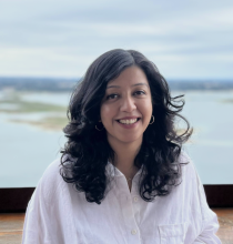 Anjali Singh wears a white button-down shirt and smiles in front of a lake with some green island patches and a cloudy blue sky.