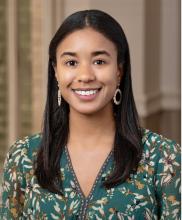 Chasitie Brown wears white hoop earrings and a turquoise blouse with a white and gold floral pattern. Behind her is a blurred indoor background.