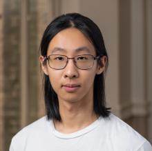 Qing Yao wears a white t-shirt and glasses. He stands in a room with brown wallpaper.