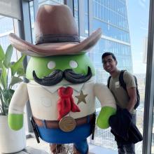 Vignesh Sella wears a black backpack and a green Oden t-shirt. He stands next to a statue of the green Android robot dressed in a cowboy costume. To the left of the robot is a plant. Behind them is a glass window overlooking Austin.