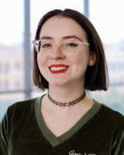 Emma Ryan wears a green blouse, a black choker necklace, and clear horn-rimmed glasses. She smiles in front of a blurred background of a floor-to-ceiling window.