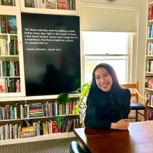 Jessie Li sits at a wooden table in a dark green sweater in front of a bookshelf. Behind her is a quote from "Jesus' Son" by Denis Johnson.