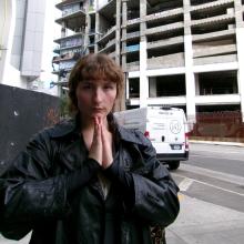 A woman wears a leather jacket and stands in front of a parking garage with her palms pressed together. Behind her a white van passes by along a concrete road.