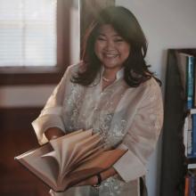 A woman wears a white blouse and flips the pages of a book. She poses in front of a bookcase and a window.