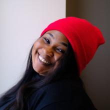 A woman wears a dark sweater and a red beanie in front of a white wall. Her head is tilted and she smiles at the camera.