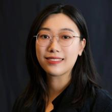 A woman wears glasses and a black blouse in front of a black backdrop.