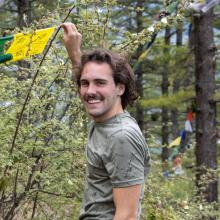 A man in a grey shirt holds up a yellow flag in front of a green bush in a forest.