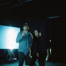Two people stand in a film studio. One sits in a director's chair.