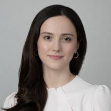 A person in a white blouse stands in front of a grey background.
