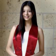 A person wears a red graduation sash and a white dress and stands in front of a brick wall.