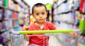 A toddler sits in a shopping cart in the middle of a grocery store.