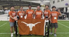 A group of students and their faculty member pose with a Longhorn flag, making the Hook'em sign and wearing orange Texas Robotics shirts, at the RoboCup competition in South Korea.