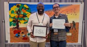 Trent Wintermeier and Donnie Sackey pose with their award certificates in front of an art piece.