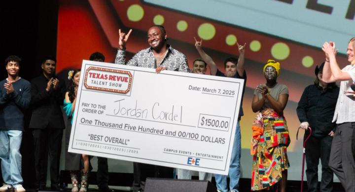 Jordan Cordel wears a black and white patterned shirt and makes the "hook em" sign onstage, surrounded by seven other people applauding, holding his giant check for one thousand, five hundred dollars from the Texas Revue.