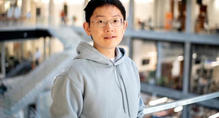 A man with black hair, grey glasses, and a grey hoodie stands in front of a metal railing. Behind him is a staircase leading to rooms lined with floor-to-ceiling windows.