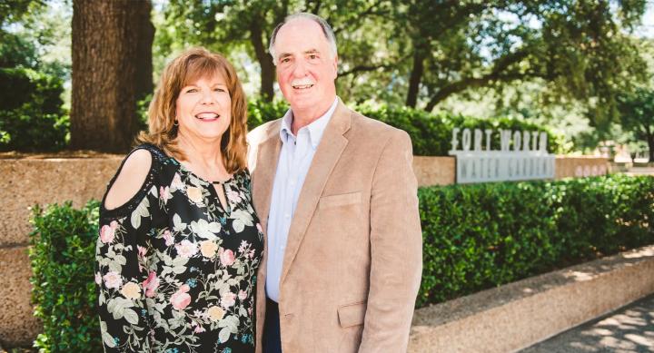 George and Barbra Polansky smile in front of a forest background. Behind them is a brown stone wall lined with a hedge, and a grey sign reading "Fort Worth Water Gardens." George wears a brown suit jacket and light blue shirt. Barbra wears a black blouse with yellow and pink flowers and green leaves.