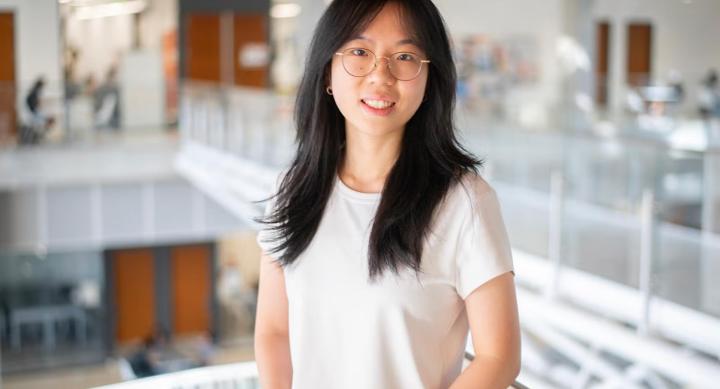 A girl with long black hair and round glasses wears a white t-shirt and stands in front of a white railing in a research laboratory.
