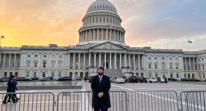 Robert Golias wears a beanie hat, a black suit, and a purple tie and stands in front of the Texas Capitol building.