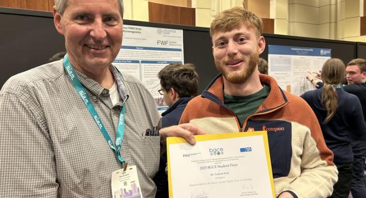 Graham Pash stands next to a professor holding his certificate for the Student Paper Prize. Behind him, a group crowds black dividers bearing research posters.