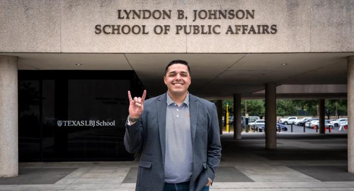 Steven Santoyo wears a grey suit jacket and makes the Hook 'em Horns hand sign in front of the Lyndon B. Johnson School of Public Affairs building.