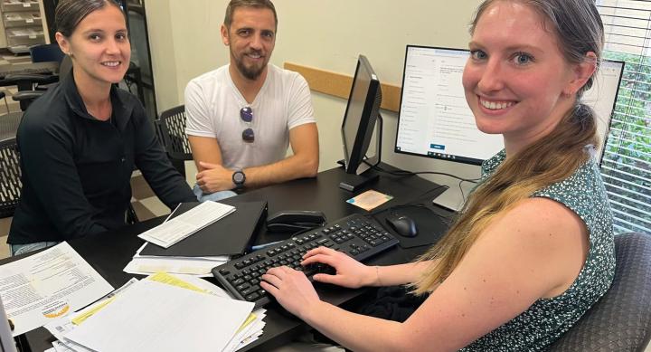 A woman sits at a desk full of tax forms and a keyboard. In front of her are a man and a woman. All three of them smile at the camera.