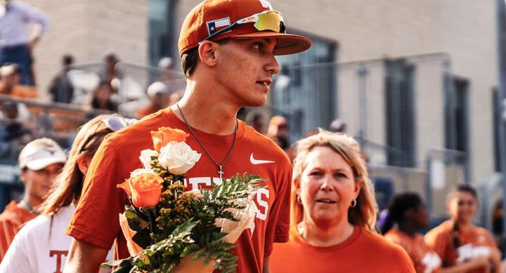 A man in an orange Texas shirt and orange Texas baseball cap holds a bouquet of orange and white flowers. Surrounding him are people in orange and white UT-Austin shirts sitting on bleachers.