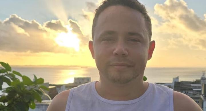 A man wears a white shirt and stands in front of a sunset over a coastal village.