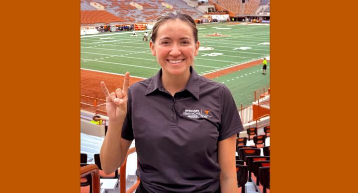 A woman in a polo shirt makes a Hook'em Horns hand gesture in front of the Longhorn football field.