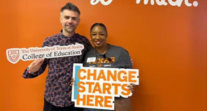 Two people stand in front of an orange wall painted with letters reading "I love coe so much." They both hold signage for the UT College of Education