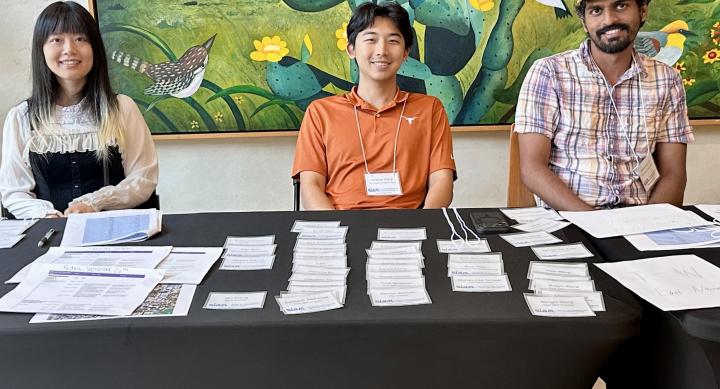 Three students wearing lanyards sit at a registration table covered in nametags and papers.