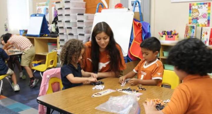 A woman helps three elementary school students as they play with a hands-on activity at a desk.