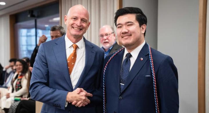 Two people in suits shake hands, one wearing graduation cords.