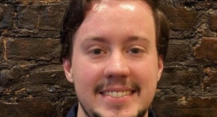 A person smiles in front of a brown brick wall.
