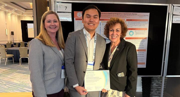 Two professors stand next to a doctoral student as they hold an award certificate in front of their poster display.