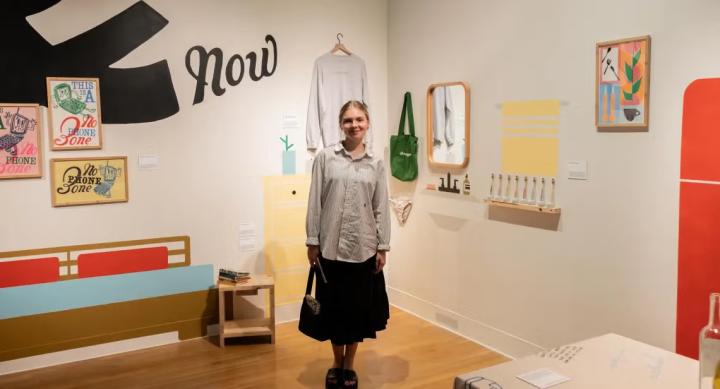 A person wears a black skirt and grey blouse and stands in an art exhibit.