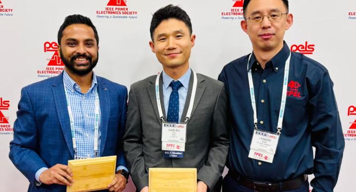 Three people wear lanyards and pose holding award plaques.