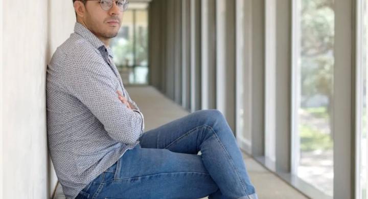 A person wears jeans and a dress shirt and sits on a padded bench in a hallway.