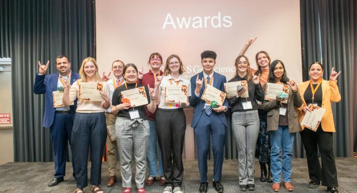 Eleven students in businesswear pose with their award certificates at the front of a conference room.