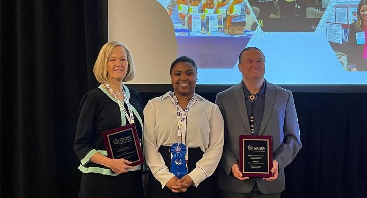Three people hold plaques and awards ribbons in front of a powerpoint presentation.