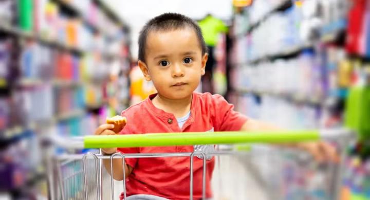 A toddler sits in a shopping cart in the middle of a grocery store.